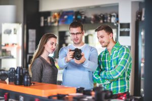 Couple is standing inside photographic equipment store and looking at digital camera that store clerk is showing to them. Lenses and other items are visible around.